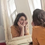 A cheerful woman smiles at her reflection in a vintage-style mirror, exuding positivity and warmth.
