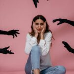 A woman looks anxious and stressed, sitting against a pink backdrop with hands reaching towards her.
