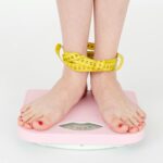High angle of crop anonymous female with centimeter on legs checking weight on scales on white background