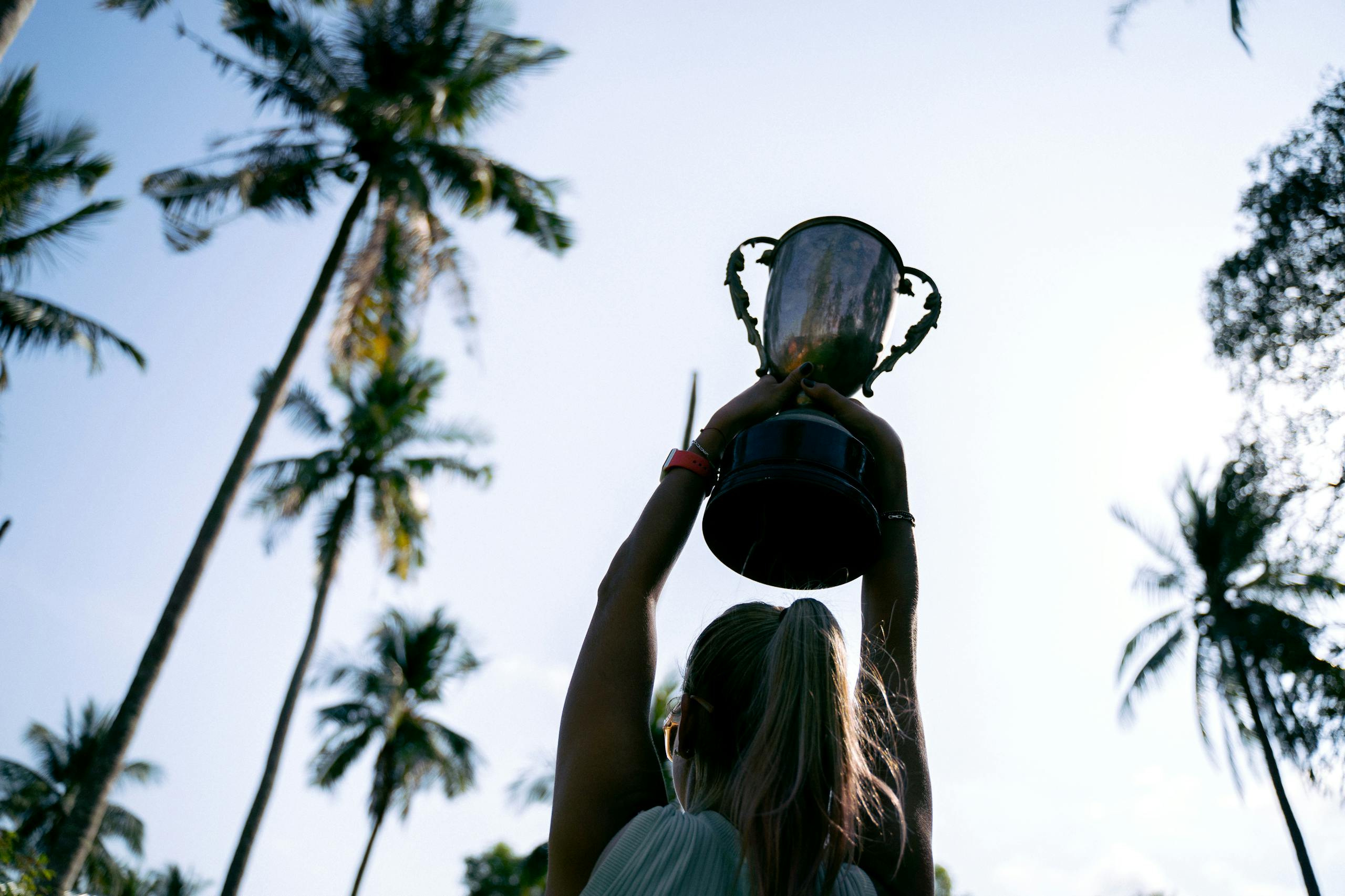 A woman lifts a trophy under palm trees, celebrating victory in nature.
