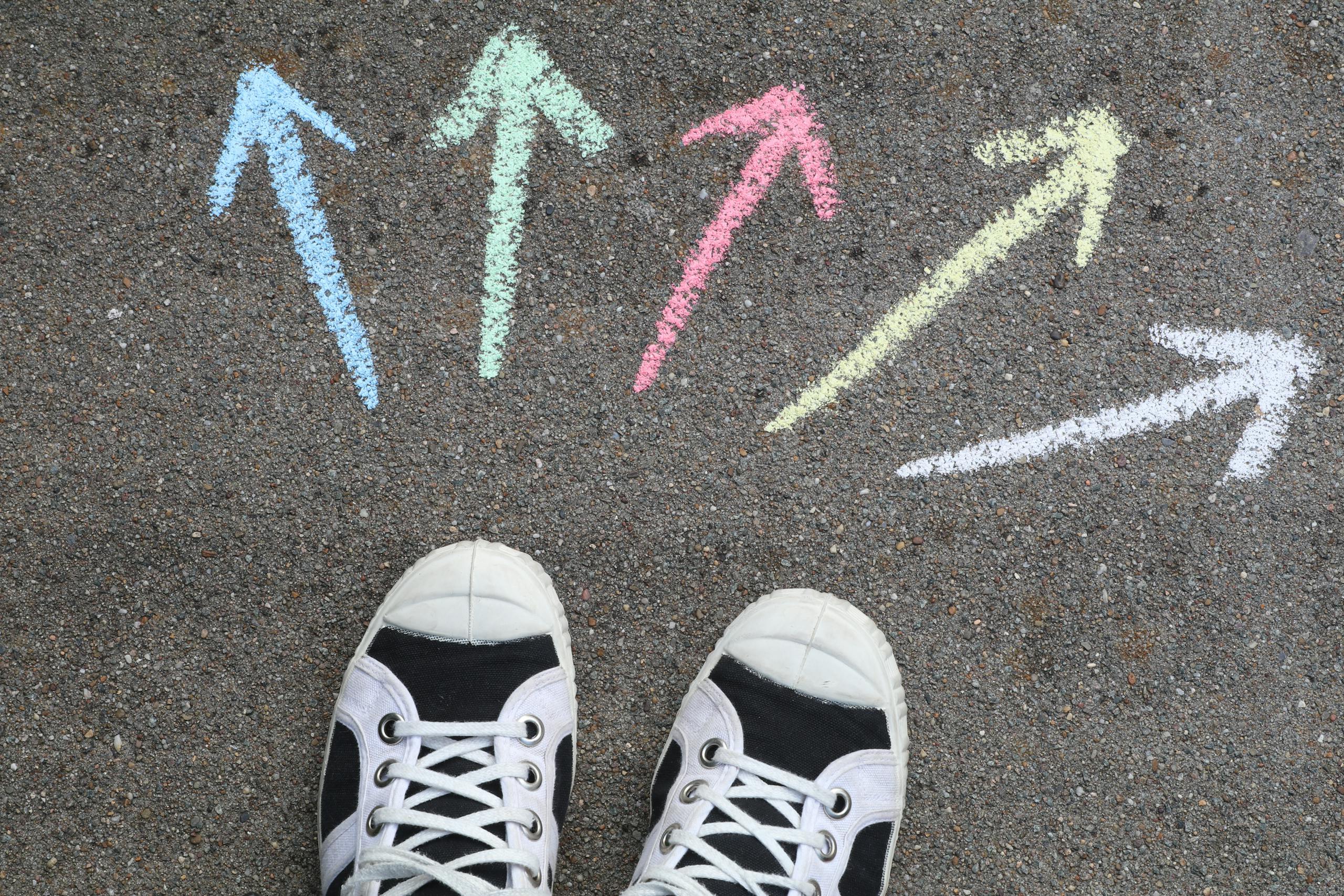 Black sneakers on pavement with colorful chalk arrows pointing in different directions.